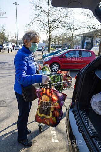 Une femme âgée portant un masque facial et des gants de caoutchouc pour se protéger pendant l'épidémie de coronavirus chargeant ses courses dans une voiture.